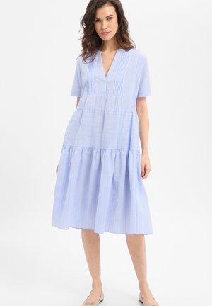 Vrouw draagt een lichtblauwe, korte mouw, knie-lange jurk met verticale strepen en een gelaagde rok, staand tegen een effen witte achtergrond.