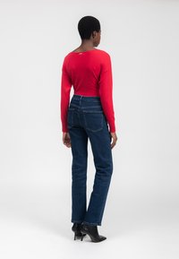 Suéter rojo de manga larga, colocado sobre unos jeans rectos de color azul oscuro. Persona lleva botas negras de tacón, vista desde atrás.