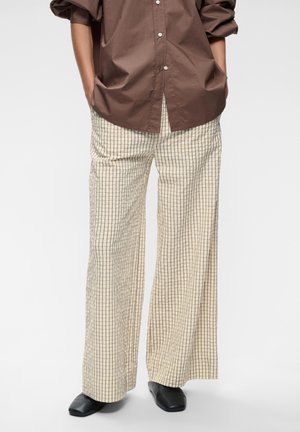 Personne portant un pantalon beige à jambes larges avec un motif quadrillé noir, une chemise marron boutonnée, les mains dans les poches et des chaussures noires à bouts carrés.