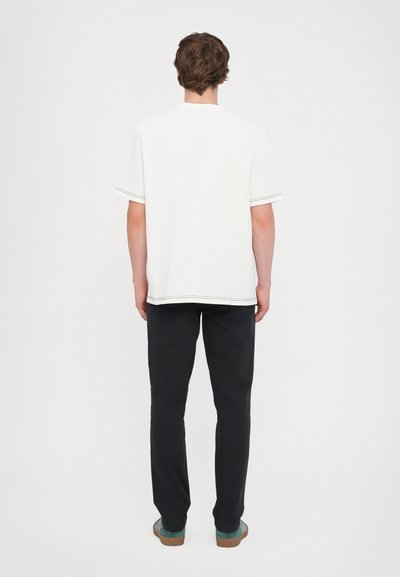 White short-sleeve t-shirt with decorative stitching, paired with dark pants and green slip-on shoes, viewed from the back.