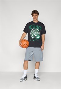 Sort T-shirt med grøn "Celtics" grafik, lysegrå shorts, holder en basketball, iført sorte og hvide sneakers med mesh-detaljer.