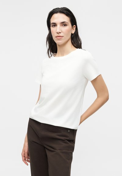 Femme aux longs cheveux foncés portant un haut blanc uni à manches courtes et un pantalon marron foncé, debout avec une main derrière le dos.