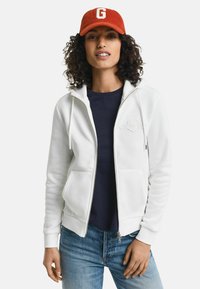 Witte zip-up hoodie met voorzakken, trekkoorden en geborduurd logo. Draag het over een marineblauwe shirt met blauwe jeans. Rode pet met de letter "G."