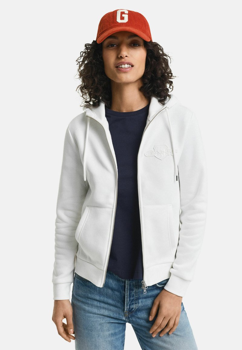 Witte zip-up hoodie met voorzakken, trekkoorden en geborduurd logo. Draag het over een marineblauwe shirt met blauwe jeans. Rode pet met de letter "G."