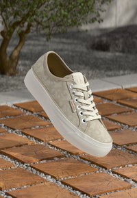 Sneaker beige di Calvin Klein con lacci, caratterizzata da un sottile motivo leopardato, suola spessa bianca e etichetta logo su una pavimentazione in legno e ciottoli all'aperto.