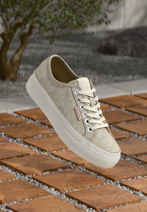 Vaaleanbeige Calvin Klein -nauhakengät, joissa on hillitty leopardikuosi, valkoinen paksu pohja ja logotunnus puuklumpilla ja kivipäällysteisellä kävelytiellä ulkona.