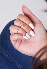 Goldene Ringe an einer Hand mit weißen Nägeln. Ein Ring ist glatt und gerade; der andere ist gedreht und hat einen kleinen rechteckigen Anhänger.