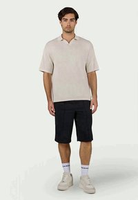 Lys beige ribbet polo skjorte med korte ermer, kombinert med mørke shorts og hvite joggesko. Detaljer inkluderer logo på sokkene.
