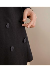 Černá bunda s texturovanou tkaninou, která má pět kulatých matně černých knoflíků a postranní rozparek, částečně zapnutá rukou.