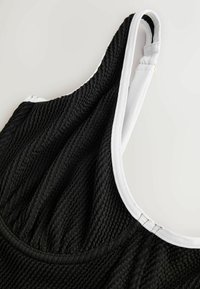 Gros plan sur un maillot de bain en tissu noir texturé avec une bordure blanche et une bretelle ajustable, sur un fond neutre.