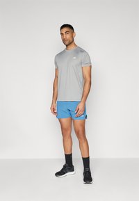 T-shirt atlético cinza de mangas curtas, combinado com calções azuis. O conjunto apresenta um pequeno logótipo e é complementado por ténis e meias pretas.