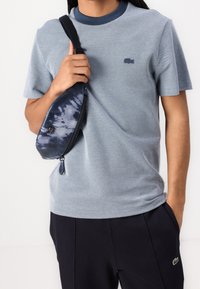 En person som bär en blå kortärmad skjorta och mörka byxor, håller en blå batikmönstrad axelväska med en hand.