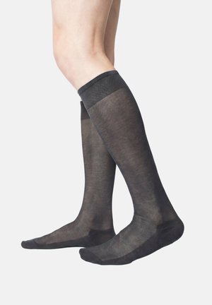 Gambe che indossano calze nere trasparenti fin sopra il ginocchio con punta, tallone e bordi rinforzati, su sfondo bianco semplice.