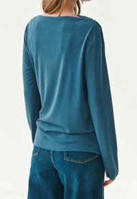 Pitkähiha, kevyt paita teallansinisessä, jossa on sileä pinta ja leveä kaula-aukko, jossa on huomaamattomat litsit hartioilla.