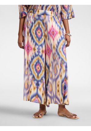 Bukser med vide ben og flerfarvet abstrakt ikat-mønster i pink, blå, lilla og sennepsfarve, båret sammen med gyldne sandaler og en matchende top.