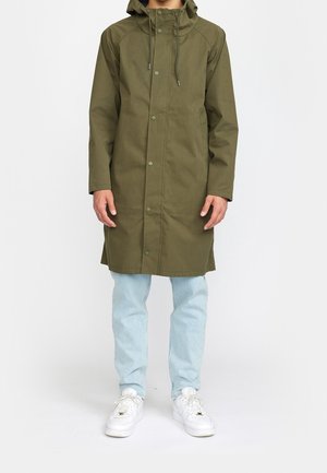 Person trägt einen olivgrünen, langen Regenmantel mit Kapuze, hellblaue Jeans und weiße Sneakers und steht vor einem einfarbigen Hintergrund.