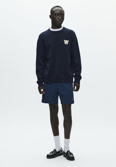 Pull en maille bleu marine avec poignets et ourlet côtelés, arborant un logo "W" blanc. Associé à un short bleu marine, des mocassins noirs et des chaussettes blanches.