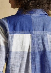 Osoba z kręconymi włosami ubrana w niebiesko-białą lnianą koszulę w kratę, widziana od tyłu, skupiająca się na górnej części pleców i kołnierzyku.