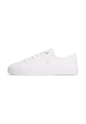White low-top sneaker with textured fabric, white laces, flat sole, and small gold logo on the side.
