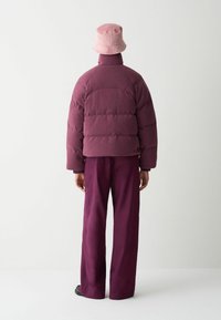 Pufjakke i burgunder, med høj krave og quiltet struktur, parret med brede, maroon bukser og en lys pink spandhat.