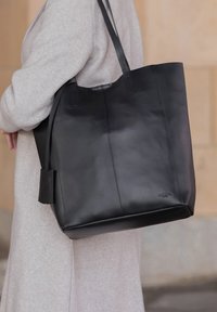 Sac fourre-tout en cuir noir avec une texture lisse, des coutures verticales et de longues anses. Dispose d'une petite étiquette sur le côté.