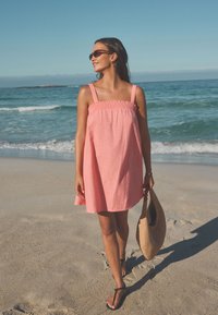 Rosa linneliknande klänning med rynkad överdel och breda axelband, kombinerad med svarta flip-flops och en ljusbrun vävd väska. Strandmiljö i bakgrunden.