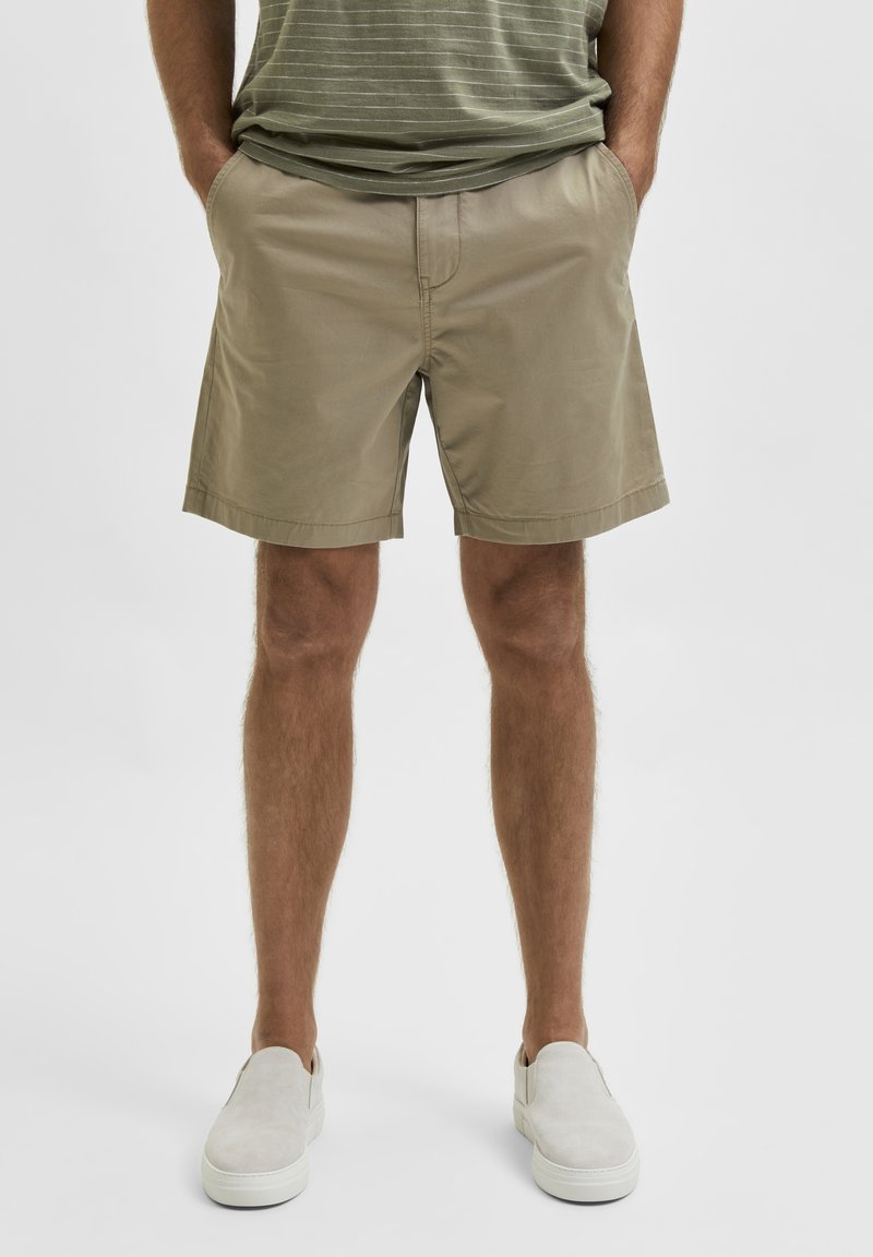 Beige bomullsshorts med framfickor, slät yta och nederkant som slutar strax ovanför knäet. Matchas med ljusgråa slip-on skor.