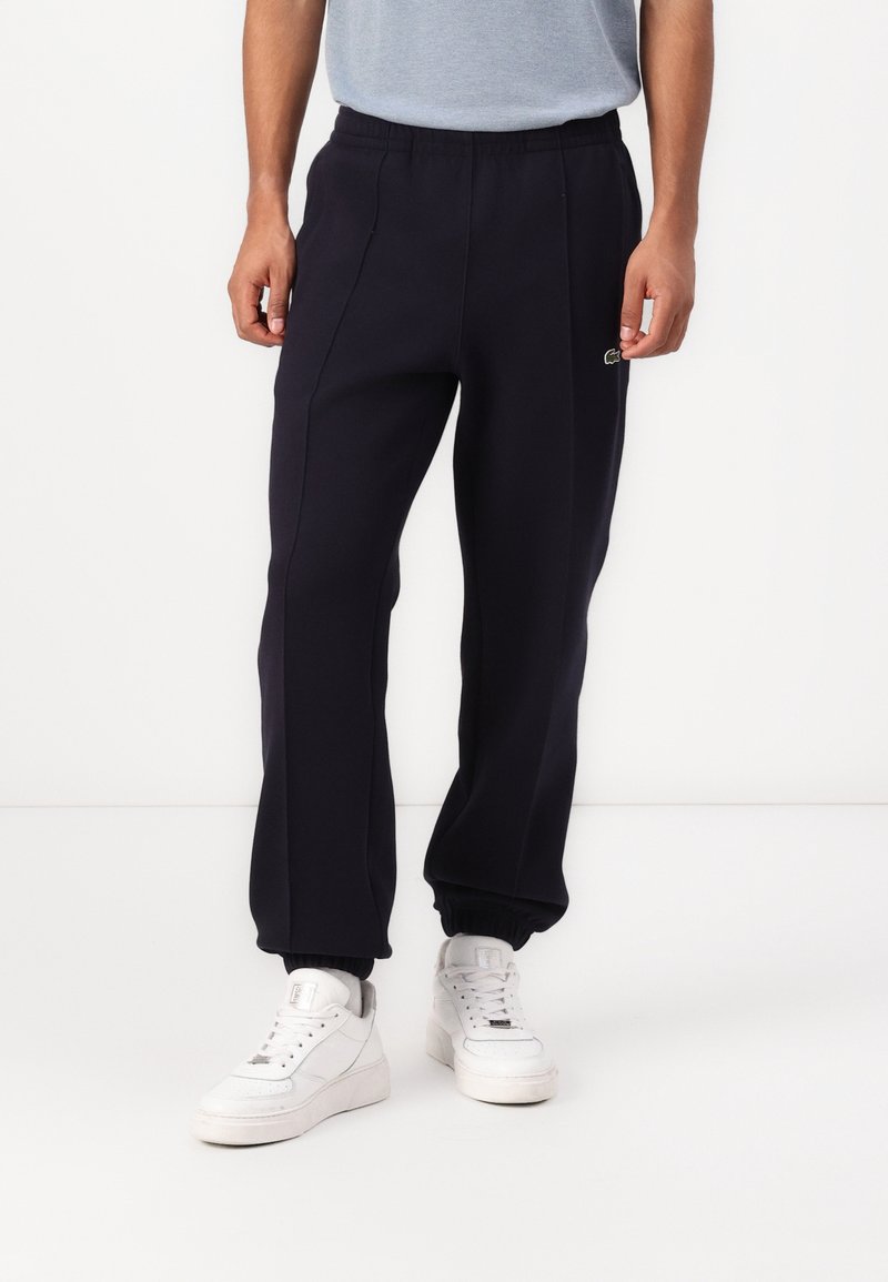 Man som bär svarta träningsoverallsbyxor med elastiska muddar och vita sneakers, stående mot en helt vit bakgrund.