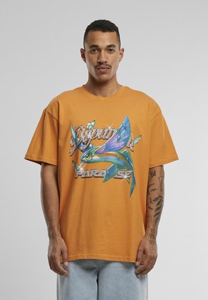 Homme aux cheveux courts portant un t-shirt orange oversize avec un motif coloré de poissons volants et un papillon, debout devant un fond uni.