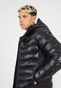 Črna puhasto jakna s horizontalnim šivom, sijočo teksturo, nastavljivo kapuco in stranskimi žepi. Vsebuje črn tekstilni panel pod pazduhami.