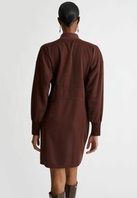 Brązowa sukienka dresowa z długimi puff sleeves, wysokim kołnierzem i dopasowaną talią. Posiada teksturowany materiał oraz elegancki, nowoczesny krój.