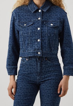 Mujer con chaqueta de mezclilla azul y jeans con un patrón geométrico a juego y botones plateados, de pie con los brazos relajados.