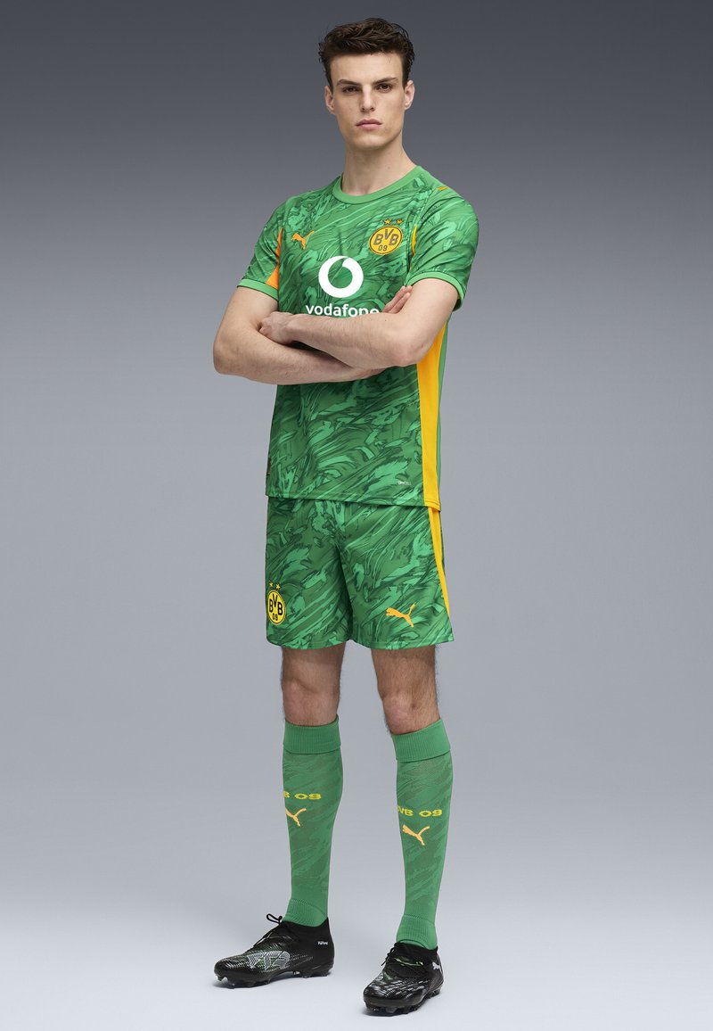 Groen voetbaltenue met een bladachtige textuur, voorzien van gele accenten. Inclusief een logo op het shirt, gecombineerd met bijpassende shorts en kniekousen.