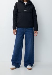 Sort hættejakke med puffer, der har en frontlomme, parret med oversized blå denimjeans. Modellen har hvide sneakers med gummisål på.