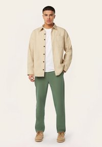 Beige overhemd met knopen over een wit T-shirt, gecombineerd met groene broek en beige schoenen. Het overhemd heeft een zak en donkere knopen.