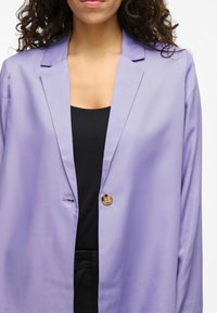 Lila blazer met een reverskraag en één knoopsluiting. Gladde textuur; gecombineerd met een zwarte top eronder.