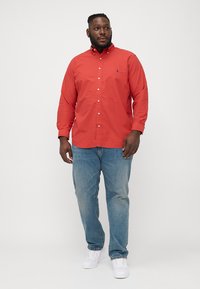 Camisa vermelha abotoada em algodão, combinada com jeans azul-claro e ténis brancos. A camisa apresenta um colarinho e um logótipo no peito.