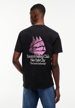 Άνδρας που φοράει μαύρο μπλουζάκι με ροζ γραφικό ιστιοφόρου και κείμενο "Tommy’s Sailing Club New York City 'The Ocean Is Our Runway'" στην πλάτη.