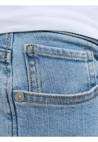 Κοντινή λήψη πίσω τσέπης μπλε τζιν με πορτοκαλί ραφή και ασημένιο κούμπωμα, φορεμένη με λευκό πουκάμισο.