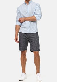 Lysblå stribet skjorte med knapper, mørke teksturerede shorts og hvide sneakers. Skjorten har oprullede ærmer; shortsene har en afslappet pasform.