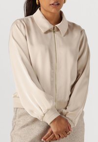 Beige satin bomberjack met een rits aan de voorkant, een overhemddetail en gathered manchetten. Textuur lichtbruine rok zichtbaar in het onderste deel van de afbeelding.