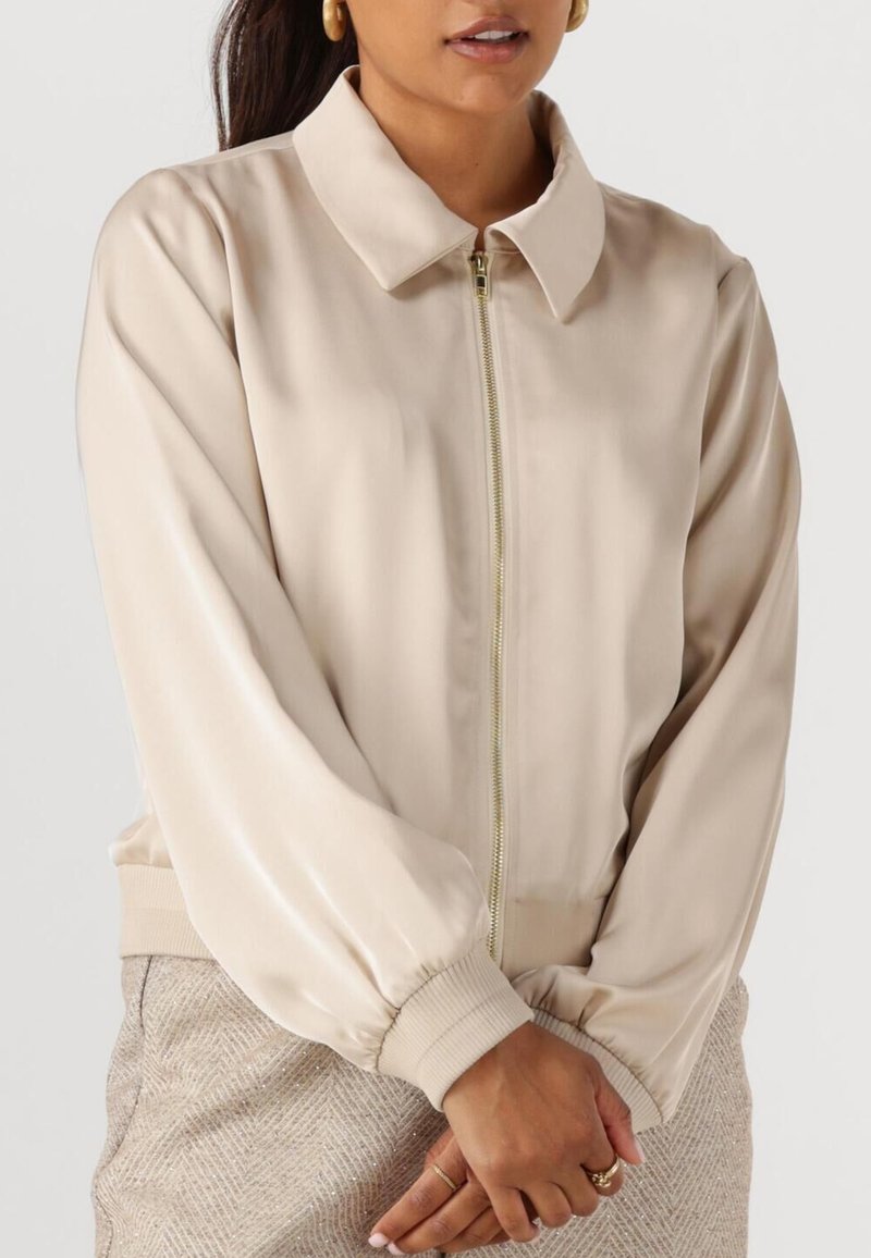 Beige satin bomberjack met een rits aan de voorkant, een overhemddetail en gathered manchetten. Textuur lichtbruine rok zichtbaar in het onderste deel van de afbeelding.