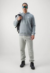 Pelēks kapučknit ar logotipu, gaiši pelēkas sporta bikses, melnas sporta kurpes un melna mugursoma. Persona valkā atstarojošas saulesbrilles.