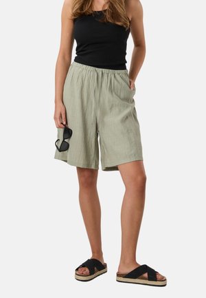 Woman wearing a black tank top, loose light olive shorts, black slide sandals, holding black sunglasses in one hand, standing against a plain background.