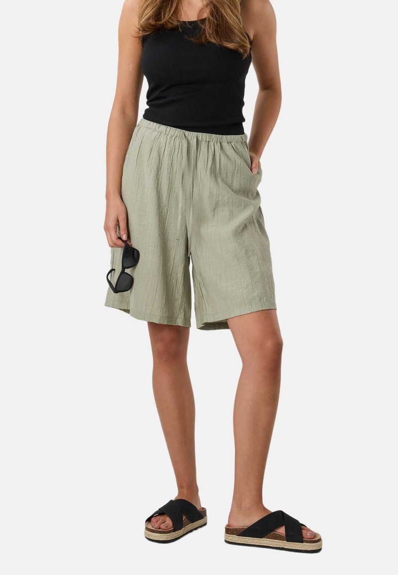 Femme portant un débardeur noir, un short ample olive clair, des sandales noires, tenant des lunettes de soleil noires dans une main, debout devant un fond uni.