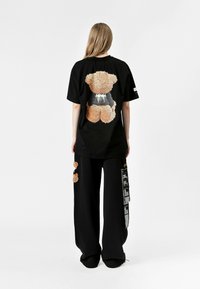 Camiseta negra de gran tamaño con un gráfico de un oso difuso en la parte posterior; combinada con pantalones anchos negros que presentan motivos de osos y detalles de texto en contraste.