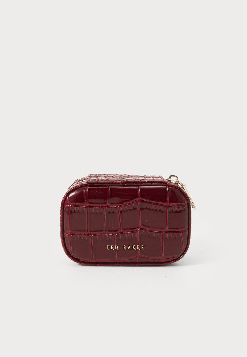 Pequeña funda rectangular con textura de cocodrilo en color burdeos, con el logotipo dorado de Ted Baker y un tirador de cremallera, sobre un fondo blanco.