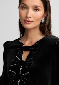 Schwarzes Samtkleid mit vier auffälligen Schleifen und glitzernden Akzenten entlang des Ausschnitts, das ein strukturiertes und detailliertes Design schafft.
