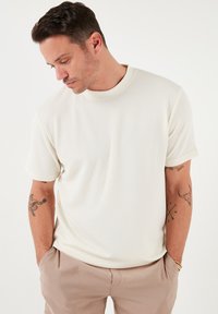 Cremefarvet ribbet kortærmet t-shirt med rund halsudskæring, parret med lys beige bukser. Har en afslappet pasform og blød tekstur.