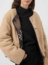 Tan faux shearling jacket with black trim; features a plaid lining in brown and white. Worn over a black turtleneck sweater.